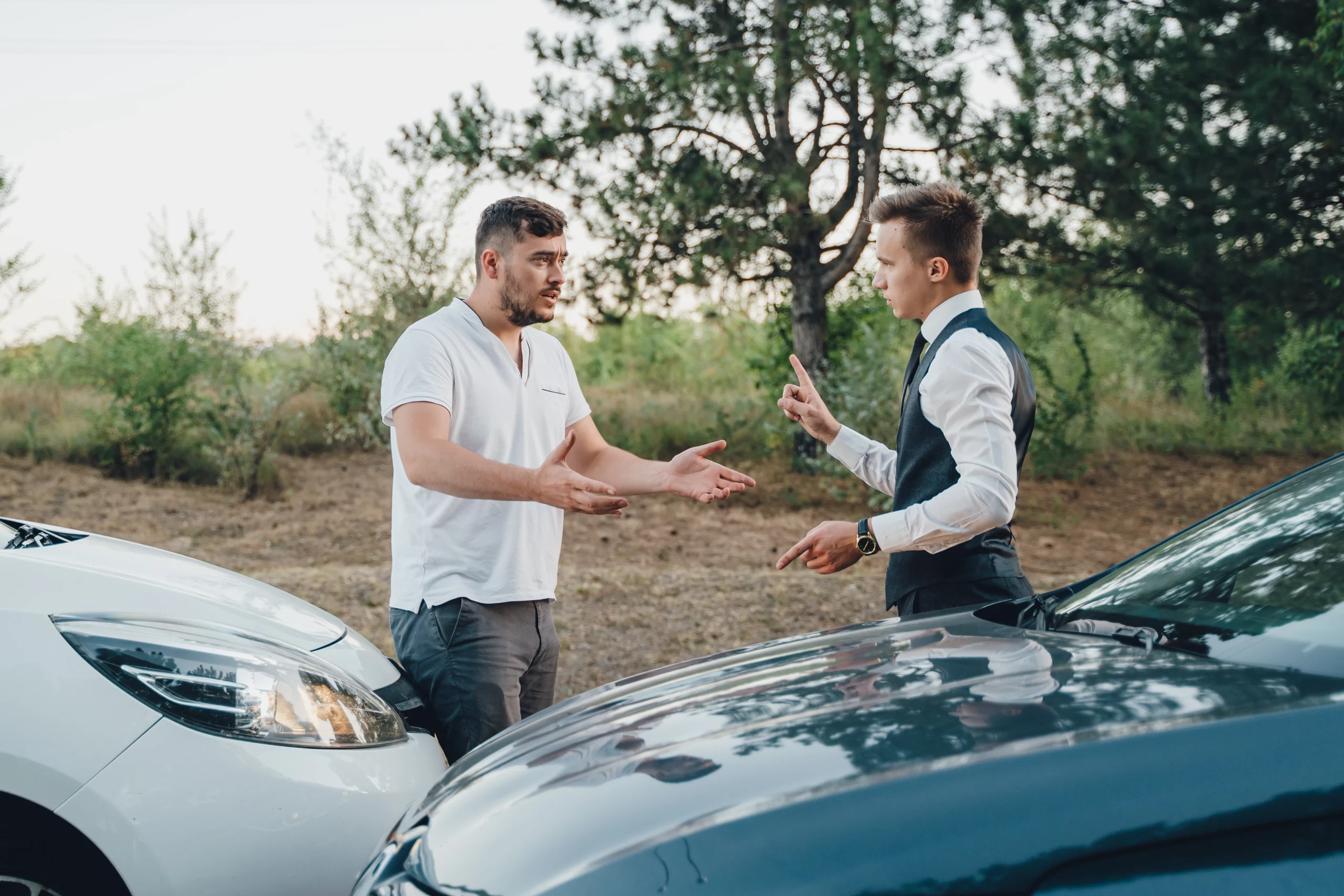 Two men standing by their cars in a forested area, gesturing and discussing after a minor car accident, showing concern and tension.