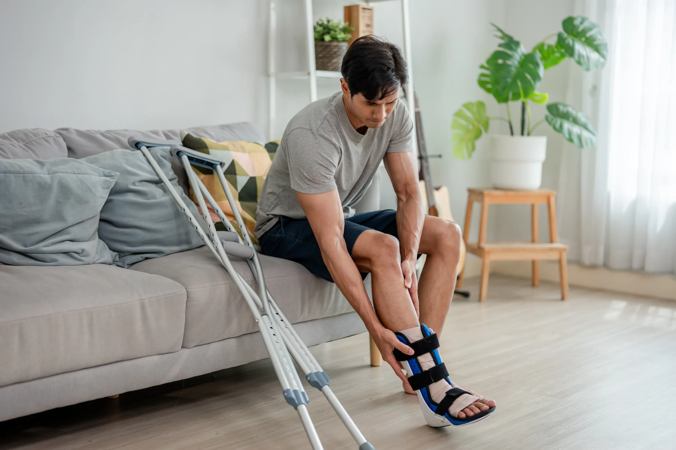 Person sitting on a couch adjusting an orthopedic boot on their injured foot, with crutches nearby in a calm home setting, illustrating recovery after an accident and factors affecting Houston personal injury case value.